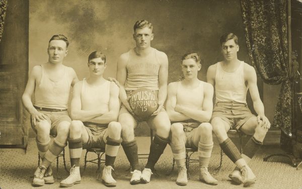 Text on the basketball reads: "Wausau Cardinals '15." Five young men are sitting on small stools for a studio portrait. The man in the center is wearing shorts and a Wisconsin tee-shirt shirt inside out and holding a basketball, and the rest of the team are wearing sleeveless tee-shirts, shorts, knee high socks and shoes. The Cardinals is the name of the Newman Catholic High School sports teams.
