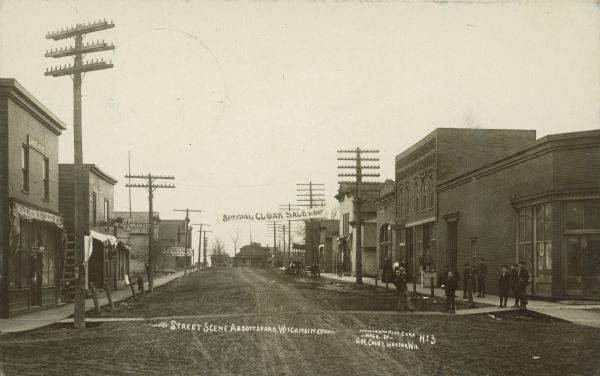 Caption reads: "Street Scene Abbottsford [<i>sic</i>], Wisconsin". A group of mainly children are standing on the boardwalk along an unpaved street lined with buildings. A horse-drawn vehicle can be seen on the right side of the street. There is a banner over the street that reads: "Special CLOAK SALE ToDAY". Signs read: "Dentist", "O.W. Bean Fruit and Confectionary" (<i>sic</i>), "Restaurant", "Clothing J.W. Shar?? Dry Goods" and "McCall's Patterns."