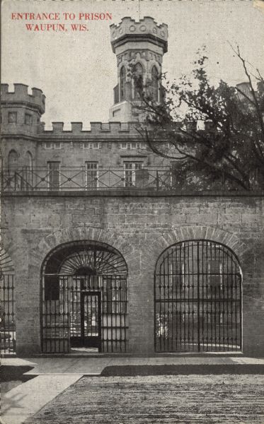 Text on front reads: "Entrance to Prison, Waupun, Wis." Main entrance of the Waupun State Prison, built in 1854. Beyond the prison gates is the tower of the Gothic Revival main building. The walls are cut limestone with wrought iron gates and grills. The prison housed adult men and women until 1933, when a separate women's prison was built.