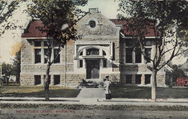 Text on front reads: "Waupun Public Library, Waupun, Wis." A woman is posing on the sidewalk in front of the library, which was built in 1904 of cut stone in the Tudor Revival Style. There are trees, a lawn and a bicycle parked near the entrance.