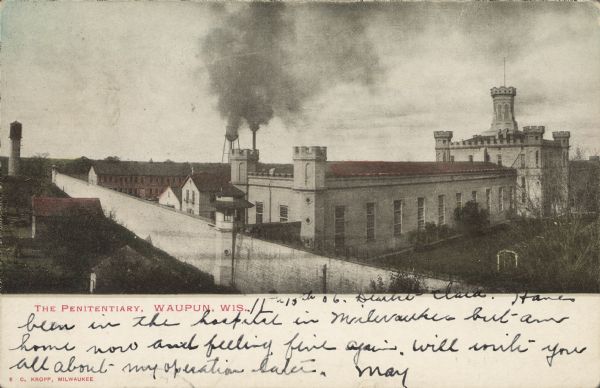 Text on front reads: "The Penitentiary, Waupun, Wis." An elevated view of Gothic Revival buildings at the prison, built in 1854 of limestone, with ornate towers. A stone wall encircles the facility. The prison housed adult men and women until 1933, when a separate women's prison was built. A water tower and smokestack can be seen on the horizon.