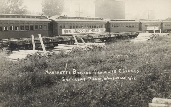 Text on front reads: "Marinette Booster Train - 12 Coaches, Settlers Picnic, Wausaukee, Wis." Five passenger railroad coaches on the tracks, one has a banner that reads: "Marinette Boosts for County Settlers Picnic." Empty train cars for transporting lumber are on the tracks in front of the coaches, and timber posts are stacked on the ground. Buildings and trees are in the background.
