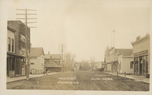 Text on front reads: "Main-Street. Wausaukee-Wis." An unpaved street lined with buildings and storefronts. A Post Office, hardware store and furniture store can  be seen. There are horse-drawn vehicles in the street and pedestrians on the sidewalks.