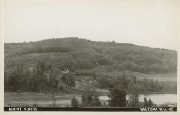 Text on front reads: "Mount Morris. Wautoma, Wis." A view of Mount Morris from above the shore of a lake or pond. A dwelling and perhaps a farm can be seen on the left. Mount Morris is six miles northeast of Wautoma. At an elevation of 1233 feet, Mount Morris is the highest point in Waushara County.