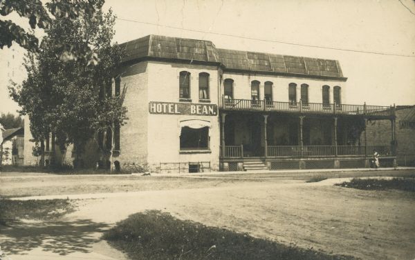 A woman on the sidewalk in front of the Hotel Bean, an Italianate style brick hotel built in 1901. It has a large covered porch with a balcony above. The hotel was demolished sometime after 1981.