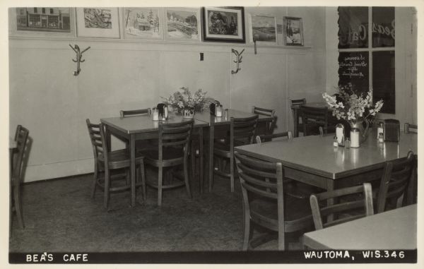Text on front reads: "Bea's Cafe, Wautoma, Wis." Tables and chairs in a restaurant with tableware and flowers on the tables. Framed paintings are displayed on an art ledge. The text: "Bea's Cafe, Serving Good Country Style Breakfasts" is painted on the window.