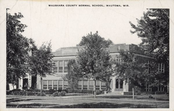 Text on front reads: "Waushara County Normal School, Wautoma, Wis." The historic name for this school was the "Waushara Training School," built in 1921 of brick in the Tudor Revival style. It is surrounded by trees, shrubs, sidewalks and a lawn. The Waushara County Department of Human Services is currently using the building.