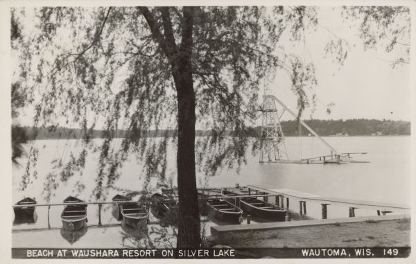 Text on front reads: "Beach at Waushara Resort on Silver Lake, Wautoma, Wis." View of a water slide in the lake from the shore. A tree and a pier with docked rowboats are in the foreground. Trees and cottages are visible on the far shore.