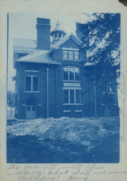 Handwritten on front: "Wauwatosa School." A cyanotype postcard of a brick, multistory school, the stone plaque reads: "Public School." Arched doorways, large windows, two chimneys and a belfry can be seen.