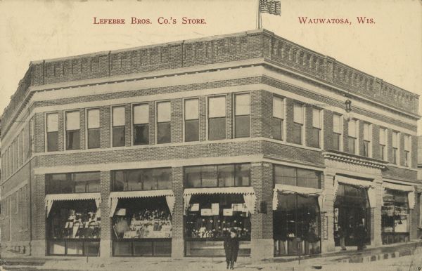 Text on front reads: "Lefebre [sic] Bros. Co.'s Store. Wauwatosa, Wis." A two story, brick building with a parapet and large display windows with transoms and awnings. The main entrance has an ornate surround. Several pedestrians are on the sidewalk. The Lefeber family owned a general store from 1896 to 1958. In 1909, three brothers opened a dry goods store. It closed in 1958 and the building burned down in 1964.