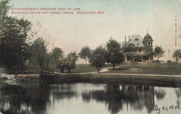 Text on front reads: "Superintendent's Residence from the Lake, Milwaukee Asylum for the Chronic Insane, Wauwatosa, Wis." A three-story house with  a porch, ornate decorations and a tower surrounded by trees. The shore of a pond, sidewalks and a bridge are in the foreground. The correct name for the institution was the "Milwaukee County Asylum for the Chronic Insane." It opened in 1880.