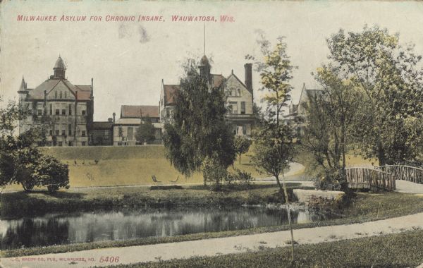 Text on front reads: "Milwaukee Asylum for Chronic Insane, Wauwatosa, Wis." Three multistory buildings with towers. A pond, trees, lawn, sidewalks and a bridge are in the foreground. The correct name for the institution was the "Milwaukee County Asylum for the Chronic Insane." It opened in 1880.