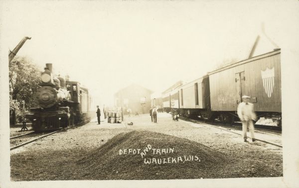Text on front reads: "Depot and Train, Wauzeka, Wis." View towards a train depot between two sets of tracks, with a locomotive on the tracks on the left, and railroad cars on the tracks on the right. People and cargo are in the center, and there is a pile of gravel in the foreground.