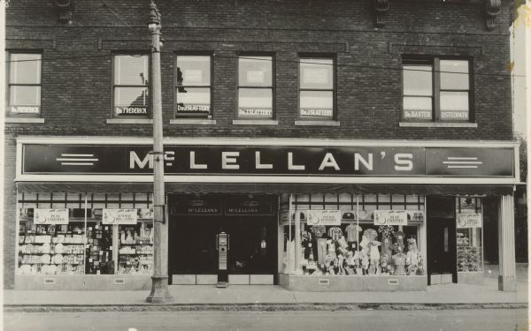 Text on reverse reads: "July 1941. This is an exterior view of our new and enlarged store at West Allis, Wis. Note the double set of doors and corner entrance. W.L. Nolan." A two-story, Neoclassical, brick building with a variety store on the first floor and doctor's offices above. The store has a sign that reads "McLellan's", three entrance doors, and large display windows full of products and merchandise. Signs read: "Summer Toys and Games", "Picnic Supplies", "Play Clothes" and "Cookies and Cakes." The doctors' names can be seen in the second story windows: "Dr. Frederick", "Dr. J. Slattery" and "Dr. Baxter, Osteopath." The building was built in 1911. Opened in the 1920s as McLellan's, renamed McCrory's in 1963, Rentmeester's M.C. Variety in 1996 and finally closed at 1:00 pm on June 21st, 2005.