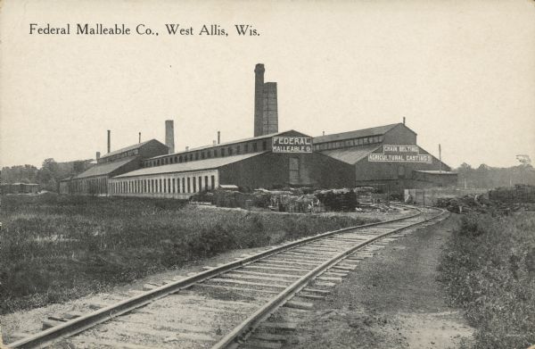 Text on front reads: "Federal Malleable Co., West Allis, Wis." A large, multi-building factory with railroad tracks leading to it. According to records, the foundry building was built of brick in 1902. Signs read: "Federal Malleable Co." and "Chain Belting Agricultural Castings." Many chimneys can be seen.