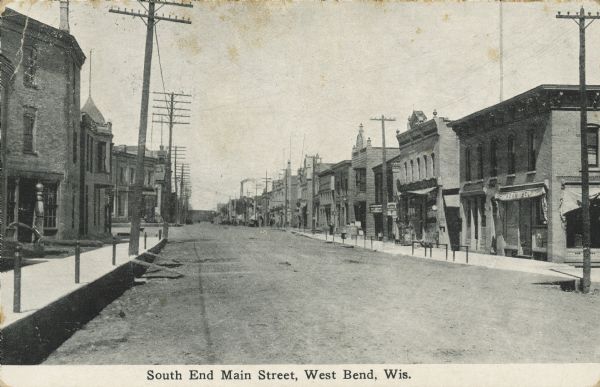 Text on front reads: "South End Main Street, West Bend, Wis." A view of an unpaved main street with sidewalks and hitching posts for horses. Buildings and storefronts are on both sides of the street. An awning on the right reads: "Adam Held."