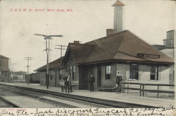 Text on front reads: "C. & N.W. Rv. Depot, West Bend, Wis." The West Bend Chicago North Western Railroad Depot was built in 1900 of clapboards in the Craftsman style. Three men are posing on the platform, one with a cart. It is listed on the State and National Register of Historic Places and has been restored.