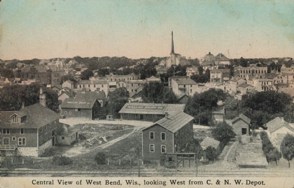 Text on front reads: "Central View of West Bend, Wis., Looking West from C. & N. W. Depot." An elevated view of the city with railroad tracks in the foreground.  A sign on two roofs reads: "Wallau Dairy Co."