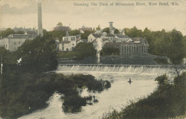 Text on front reads: "Showing the Dam of the Milwaukee River, West Bend, Wis." Elevated view of the West Bend dam with a brewery on the left in the background. The Milwaukee River has wooded banks and buildings of the city are in the background.