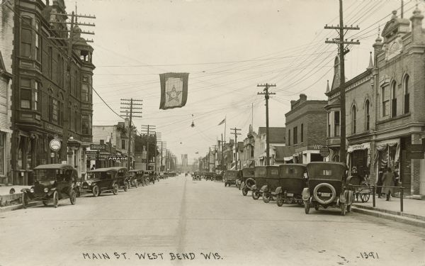 Text on front reads: "Main St. West Bend, Wis." View down center of a busy street with businesses, storefronts, pedestrians and many parked automobiles. Signs read: "Dentist", "Drugs, Ice Cream, Souvenirs, Candies, Stationary, Cigars, Kodak", "Central Auto Co. Garage" and "West Bend Post Office, Wisconsin." An American Flag and a banner with a star are hanging over the street on wires.
