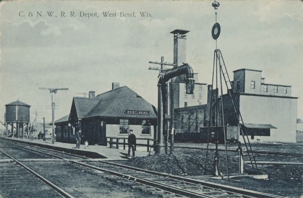 Text on front reads: "C. & N.W. R.R. Depot, West Bend, Wis." View across two sets of railroad tracks towards the platform where two men and a dog are standing. Also near the platform are a water pump, water tower, train signals and a horse-drawn wagon. The West Bend Chicago North Western Railroad Depot was built in 1900 of clapboards in the Craftsman style. It is listed on the State and National Register of Historic Places and has been restored.