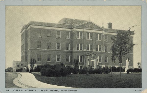 Text on front reads: "St. Joseph Hospital, West Bend, Wisconsin." Built of brick in the Neoclassical style. A statue is on the lawn in front of the entrance. The hospital opened in 1930 and was staffed by the Sisters of the Divine Savior. A new hospital was built in 2005.