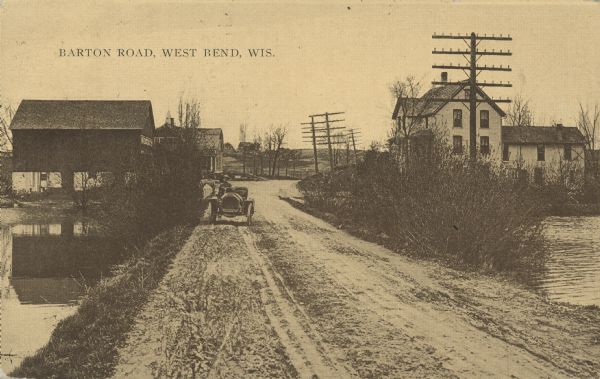 Text on front reads: "Barton Road, West Bend, Wis." A man is driving an automobile on a muddy, unpaved road with a pond on both sides. Two farm buildings are on the left and a home is on the right. There are tall shrubs on the banks of the pond.