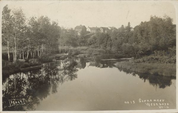 Text on front reads: "Scene Near Westboro, Wis." Water scene with a boat at the shore on the left. Trees, shrubs and foliage fill the landscape along the shoreline. In the distance are buildings on a hill.