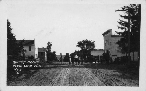 Text on front reads: "Street Scene West-Lima, Wis." A group of six children, of various ages, pose in the middle of an unpaved street. An automobile and a horse-drawn wagon are in front of the storefront on the right. Buildings and trees are on both sides of the road.