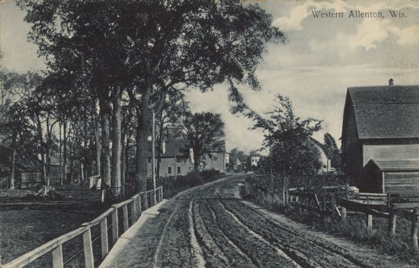 Text on front reads: "Western Allenton, Wis." An unpaved road with deep ruts, fences and trees. There are farm buildings on the right, a pasture on the left and buildings of the town in the distance.