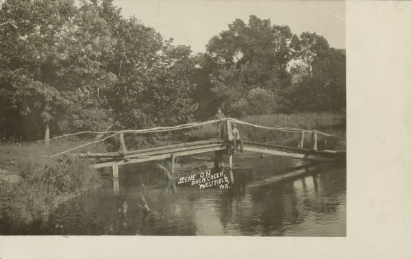 Text on front reads: "Scene on Buck Creek, Westfield, Wis." A seated man and a standing boy pose on a wooden bridge over Buck Creek. The bridge has railings made of tree branches. Trees are in the background.