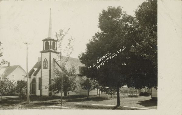 Text on front reads: "M.E. Church, Westfield, Wis." A Methodist Episcopal Church with a belfry, built in 1865 of clapboard in the Gothic revival style. There are buildings and trees on both sides.