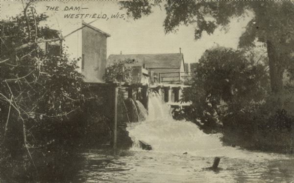 Text on front reads: "The Dam – Westfield, Wis." Water spills over the dam into the river, and buildings are behind and above the dam. There are trees on both shorelines.