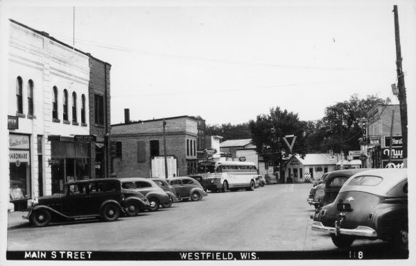 Text on front reads: "Main Street, Westfield, Wis." Many automobiles and a Greyhound Bus are diagonally parked on Main Street. The destination sign on the bus is "Madison." Many businesses are on both sides of the street with "Westfield Milling & Electric Light Co." at the far end. Signs read: "Hamilton Bros. Hardware", "JHO. Conant, Lawyer", "Westfield State Bank", "Post Office, Westfield, Wisc.", "Rexall Drugs", Millers High Life Beer", Blatz Beer", "Home Restaurant", Sun-Proof Paint, G.A. Alexander Hardware" and Murphy's Bar." There is also a large sign titled: "Roll of Honor" with a list of names.