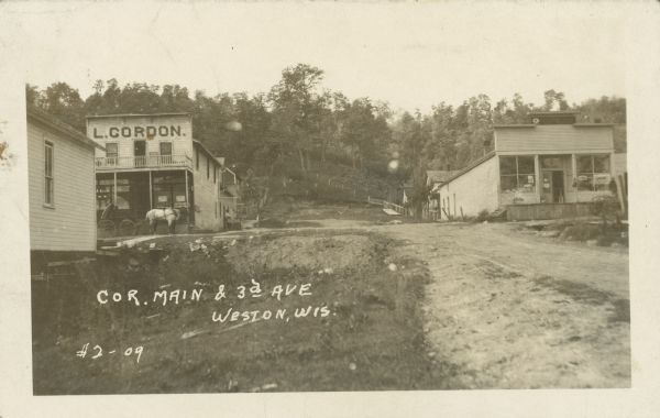 Text on front reads: "Cor. Main & 3d Ave., Weston, Wis." Three buildings at an intersection of two unpaved streets. On the left is a store with a sign that reads: "L. Gordon", a horse and buggy with passengers is in front and a man is standing long the side. On the right is a shoe store. Between the stores in the background are trees and fences on a hill.