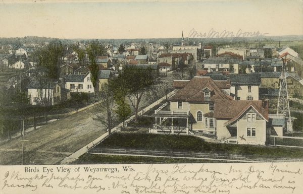Text on front reads: "Birds Eye View of Weyauwega, Wis." Elevated view of the town with a neighborhood in the foreground. A street runs diagonally towards the downtown area. There is a windmill on the far right in the foreground, behind a large house.