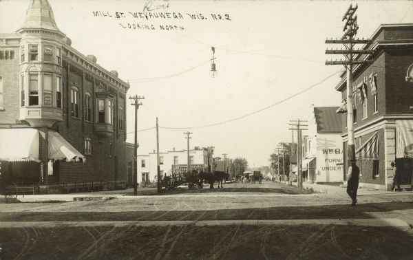 Text on front reads: "Mill St., Weyauwega, Wis., Looking North." An unpaved street with horse drawn vehicles and pedestrians. Storefronts and sidewalks line the street. Signs read: "Doctor Jones Office", "Exchange Block", "Bennet & Myers Furniture", "Wm Ba[????] Furniture Undertaker."