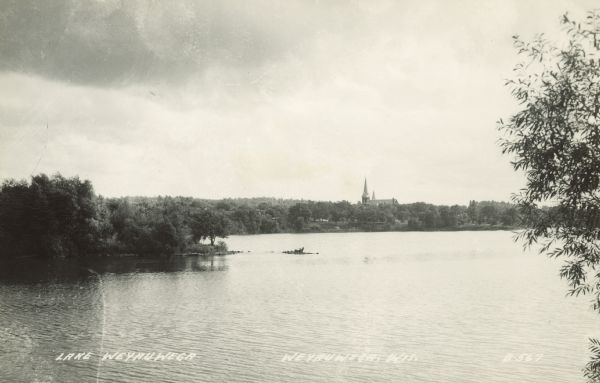 Text on front reads: "Lake Weyauwega, Weyauwega, Wis." A lake created by a dam on the Waupaca River with a wooded shoreline. The far shore is in the distance, with St. Peter's Evangelical Lutheran Church above the trees in the center. It was built in 1909 of brick in the Gothic Revival style.