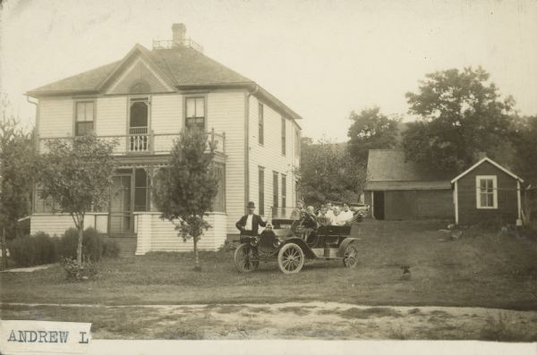 Partial label on front reads: "Andrew L." Handwritten on reverse: "Residence of Andrew Lien, Wheeler, Wis." A family poses with their automobile next to their two-story, clapboard home. A man and woman are standing on the ground on the passenger side, a young boy is in the driver's seat, and four people are in the back seat. A dog and more buildings are on the right. The license plate number is 14452W.