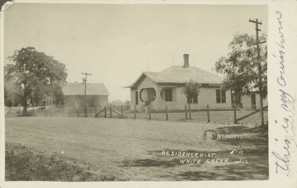 Text on front reads: "Residence, Dist. White Creek, Wis." Handwritten on front: "This is my cousin's house." A home with oblong-shaped openings on the porch. The exterior of the porch is covered with strings, presumably for vines to grow on. More buildings can be seen behind the house on the right, and other buildings are to the left among trees. A wire and post fence is along the unpaved street.