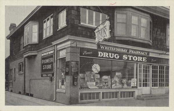 Text on reverse reads: "Whitefish Bay Pharmacy, Inc. N. Lake Drive & Silver Spring Road. Whitefish Bay, Wis. Tel. Edgewood 7680, 7681, 7682." A corner Drug Store with large display windows, built of brick with a wood shingled second story. The windows are filled with merchandise. Many signs read: "Drugs", "Whitefishbay Pharmacy", "Pioneer Store", "Soda Grill" and "Drug Store."