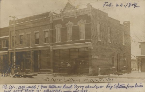 Text on front reads: "Post Office Building, Whitehall, Wis." Known as the "Post Office Block," the building was constructed in 1898 by postmaster Christopher E. Scott, one of the first settlers of Whitehall. A woman and two men are standing near the doorway and a sled and horses are on the left. It is built of brick in the Italianate style and held the post office and other businesses. Signs read: "Post Office", "Dr. L.N. Larson, Veterinarian", "Saloon" and "Hotel Best." Today, the building is a restaurant with a cafe next door.
