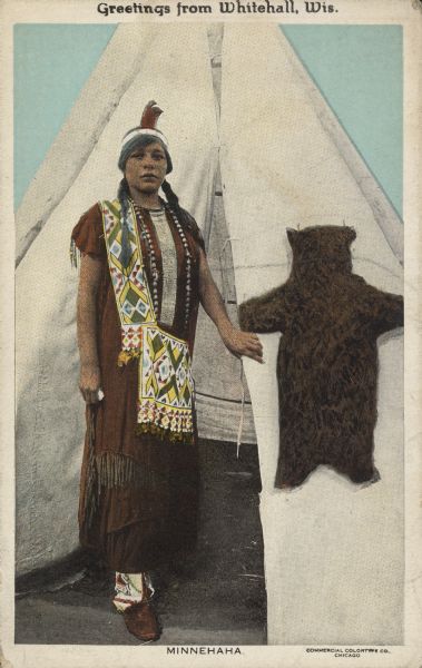 Text on front reads: "Greetings from Whitehall, Wis. Minnehaha." A Native American woman in indigenous dress, standing in the entrance to a tipi. A small bear pelt is pinned to the tipi on the right. Minnehaha is a fictional Native American woman featured in literature and art.