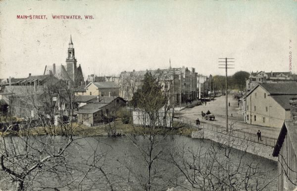 Text on front reads: "Main Street, Whitewater, Wis." Elevated view over water towards the downtown area, with buildings and storefronts along Main Street which curves to the right. A horse-drawn wagon and pedestrians are in the street and on the sidewalks. The building with the tower on the left is the Old City Hall, built in 1900 and demolished in 1971. The water in the foreground may be Cravath Lake.