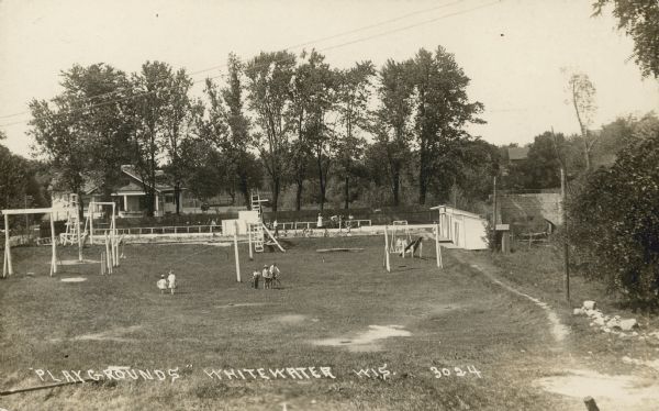Text on front reads: "'Playgrounds,' Whitewater, Wis." Two girls, and three boys with a bicycle are in a grass covered playground with swings, slides, seesaws, basketball hoops and small buildings. A pool is located at the back of the park, full of swimmers, with a water slide. A low rail fence is at the back of the pool, with several groups of people and a horse in an area of grass. Beyond is a high board fence that separates the park from a house with a large yard full of trees.