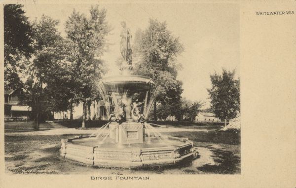 Text on front reads: "Birge Fountain. Whitewater, Wis." The Neoclassical-style Birge Fountain, located in Flat Iron Park. Mythical figures decorate the foot of the structure and a woman pouring water from a basket tops the fountain. Below her are dolphin-riding cherubs. Beyond the fountain is a street with homes and trees. Today, the fountain stands in front of the Whitewater Chamber of Commerce Building. From the Property Record on the Wisconsin Historical Society website: "the Birge Fountain, which was donated to the city by Julius Birge in 1903. The fountain is 17.5 feet high and 9 feet in diameter. Julius required that the fountain be placed on the site of the little brick school where he learned to read and write."