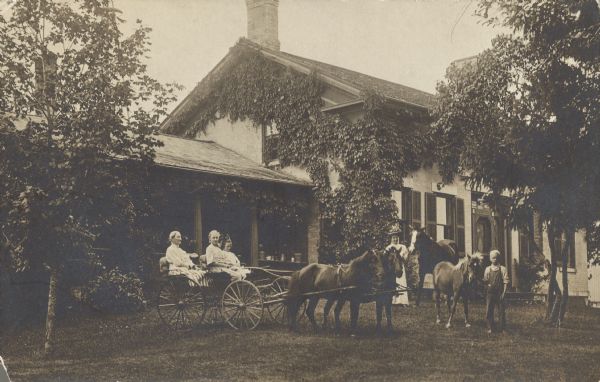 Handwritten on reverse: "Eva Kelly's old house in Hebron." A family in front of their house, with people in a horse-drawn buggy, and a boy and a woman standing on the lawn with horses. The house is two stories and built of brick, with stone lintels above the windows and doors, and shutters on the windows. Vines are growing on the sides of the house, which is surrounded by a lawn and trees.
