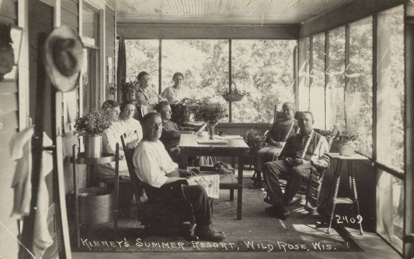 Caption reads: "Kinney's Summer Resort, Wild Rose, Wis." A group of adults, seated and standing, on a screened porch at a resort. The man in the foreground is reading "The Saturday Evening Post". The porch is furnished with tables and chairs, and decorated with flowers and plants. Trees can be seen through the screens.