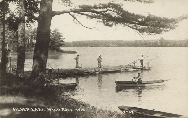 Text on front reads: "Silver Lake, Wild Rose, Wis." View from shoreline towards three men standing on an earthen pier, two fishing and one watching. Another man is rowing a boat near the shore where two more boats are moored. Large trees overhang the lake, and the shoreline is wooded, and with a marshy area on the left. Cottages are along the far shore.