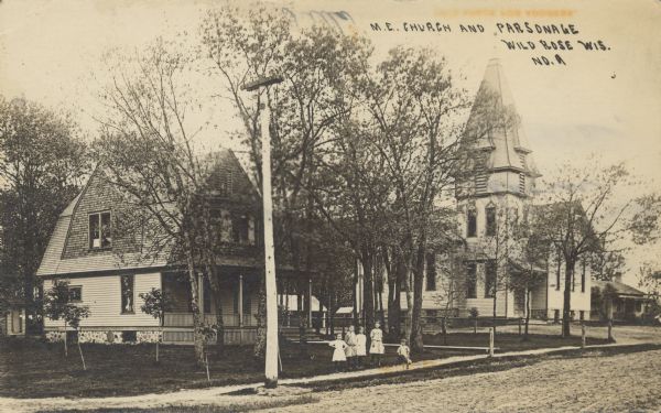 Text on front reads: "M.E. Church and Parsonage, Wild Rose, Wis." View across street towards four girls and a boy posing on the sidewalk in front of the parsonage, next to the church, which is on the right. Both buildings are built of clapboard with rubble stone foundations. The street is unpaved and lined with trees. More buildings are in the background on the right.